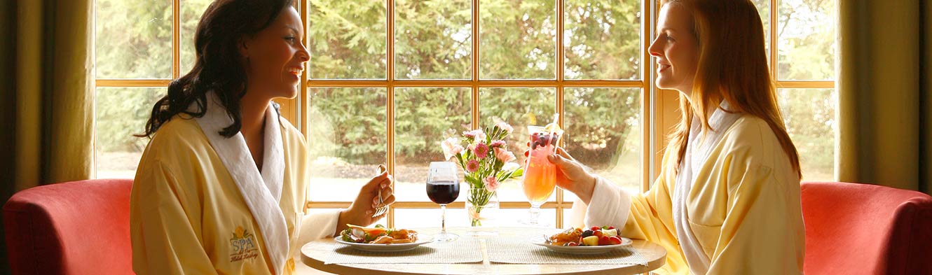 Two women in complimentary white Spa robes, sitting at a table by the window enjoying meals and drinks.